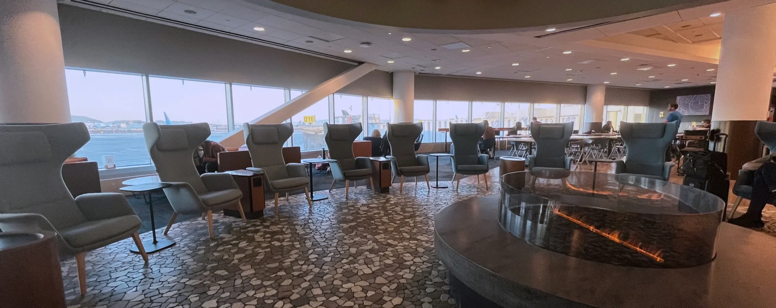 Airport lounge with wingback chairs, a round fireplace, and tarmac views